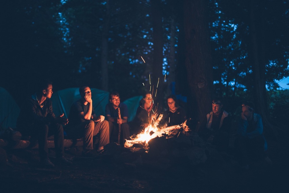 grupo de personas alrededor de una hoguera en un bosque disfrutando de la noche y con tiendas de campañas atrás