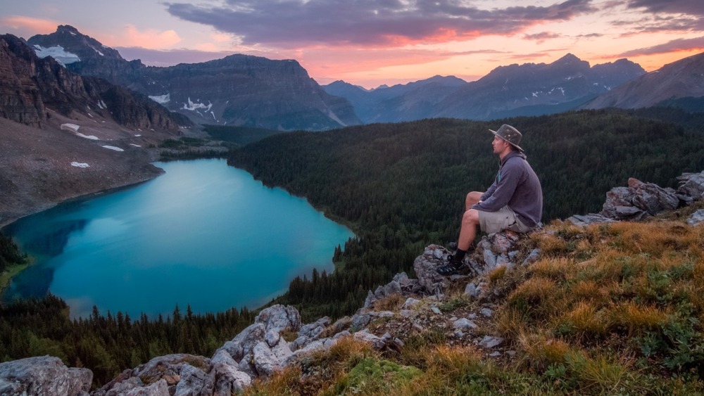 Atardecer en un paisaje montañoso y un lago. Hombre con vestimenta de aventurero sentado en unas rocas disfrutando del momento.