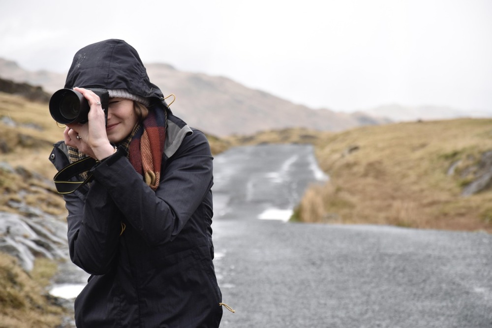 mujer mirando por el visor de una cámara y realizando una fotografía muy abrigada. Camino de grava y paisaje montañoso, nublado y frío.