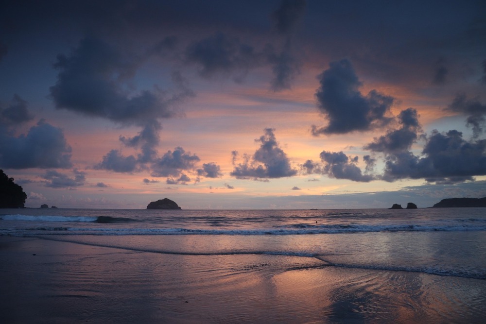 Atardecer en la playa con cielo violeta y nubes oscuras, reflejos cálidos en la arena mojada y formaciones rocosas al fondo
